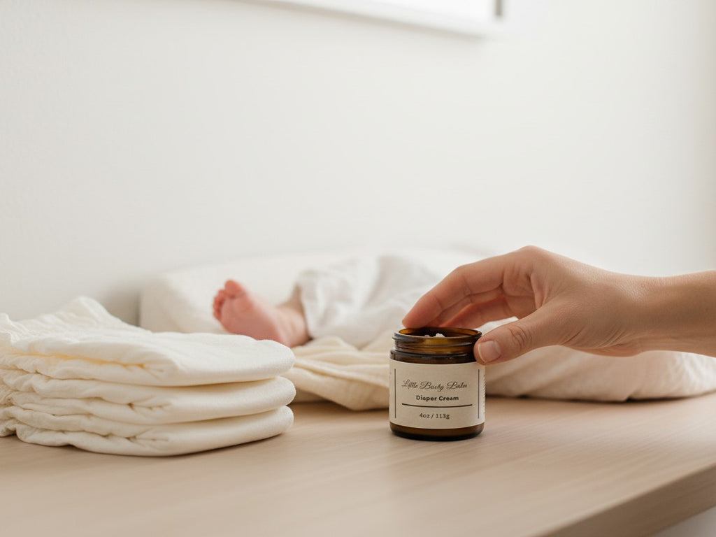 Fjell Dekor Little Booty Diaper Balm in a 4oz amber glass jar held by a hand near a stack of cloth diapers
